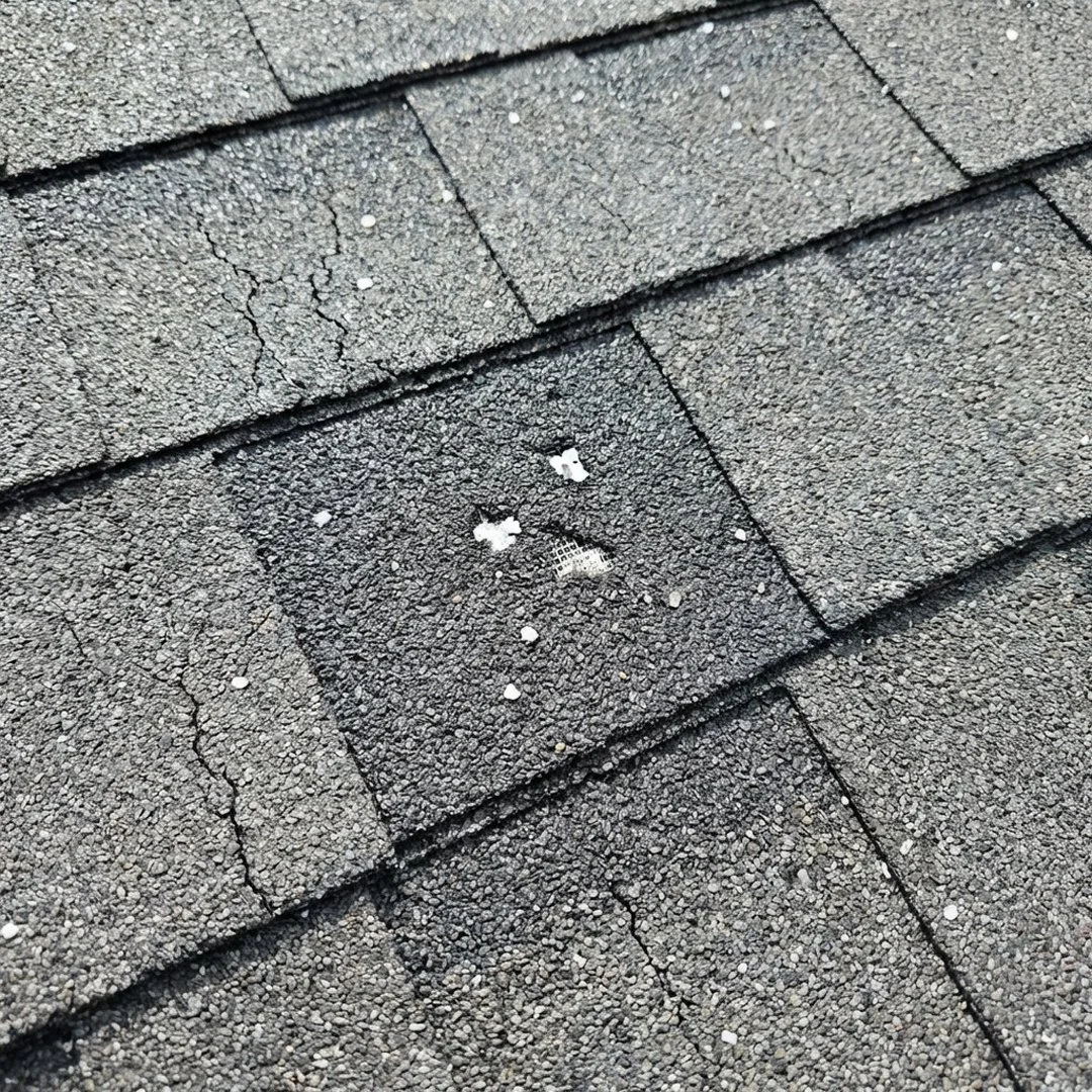 Close-up of asphalt shingles on a roof, showing a small patch of damaged shingles with cracked and missing pieces.