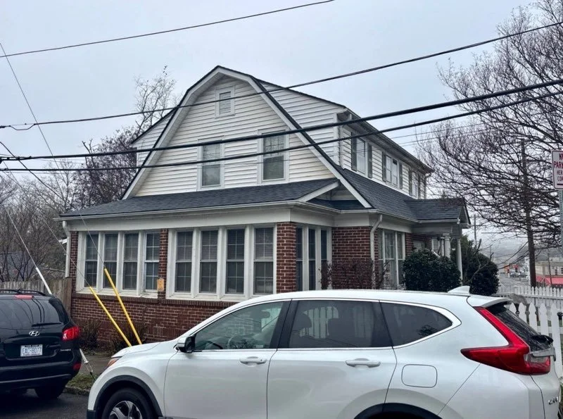 Two cars parked in front of a two-story house with white siding, brick base, and a front porch. Power lines are visible above the house.