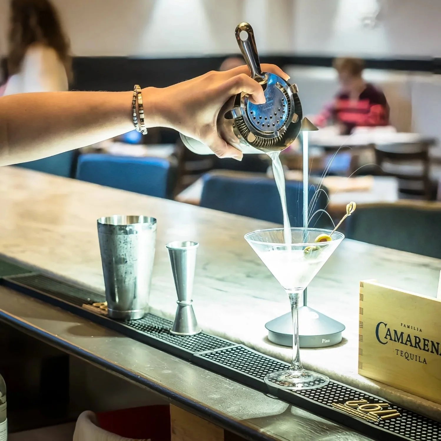 Person pouring a cocktail into a martini glass at a bar, with bar tools and a Camaren tequila sign on the counter.