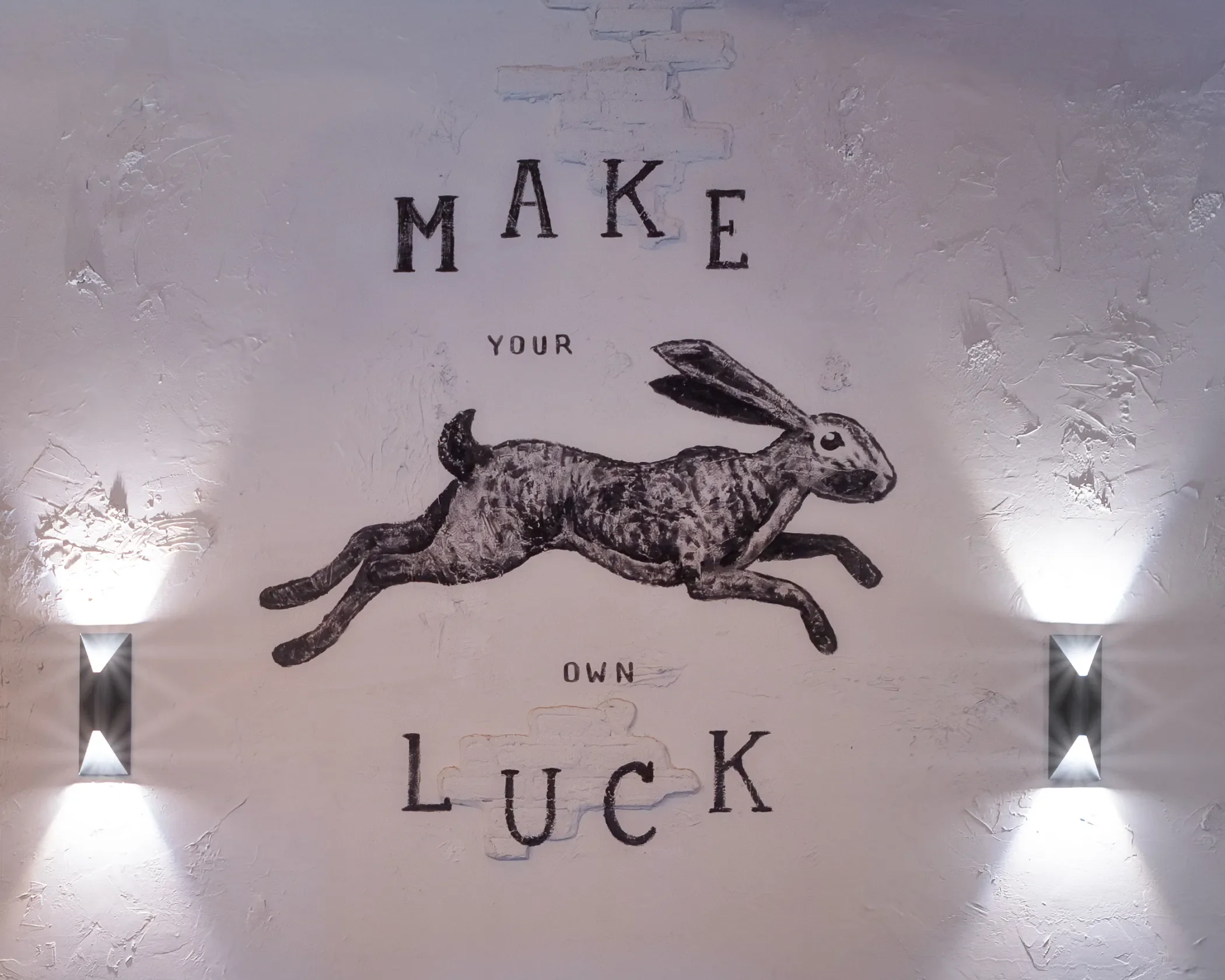 Wall art with the phrase "Make your own luck" and an illustration of a rabbit with a dog’s body, illuminated by two wall lights.