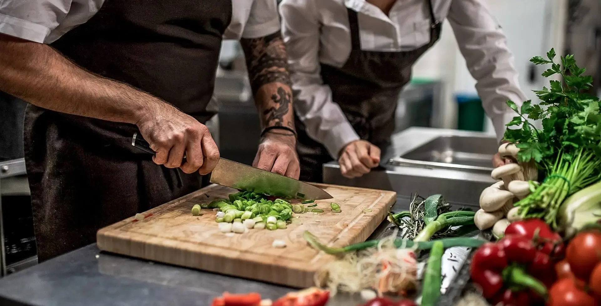 Two people chopping green onions on a wooden cutting board in a kitchen with fresh vegetables on the counter.