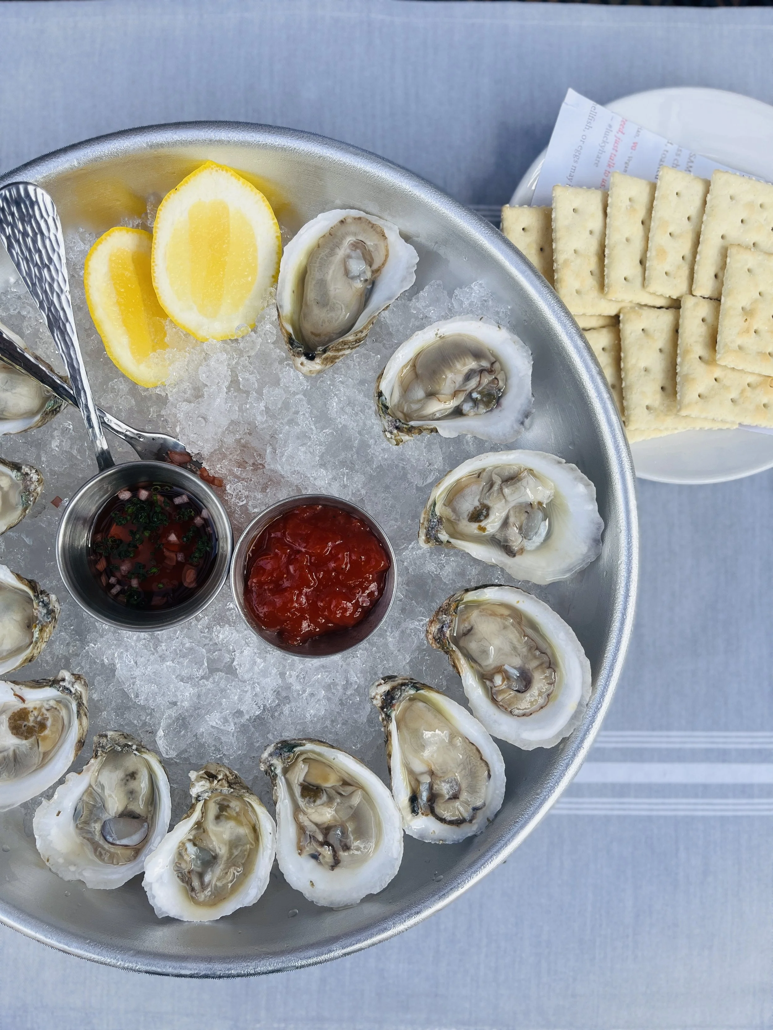 A metal tray of raw oysters on ice with lemon wedges, two small containers of cocktail sauces, and a plate of saltine crackers.