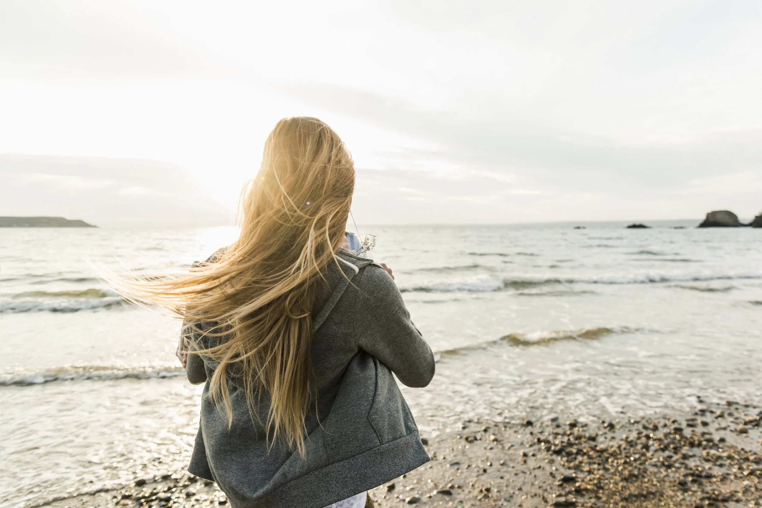 A woman with long blonde hair looking out at the ocean waves at a beach during sunset or sunrise.