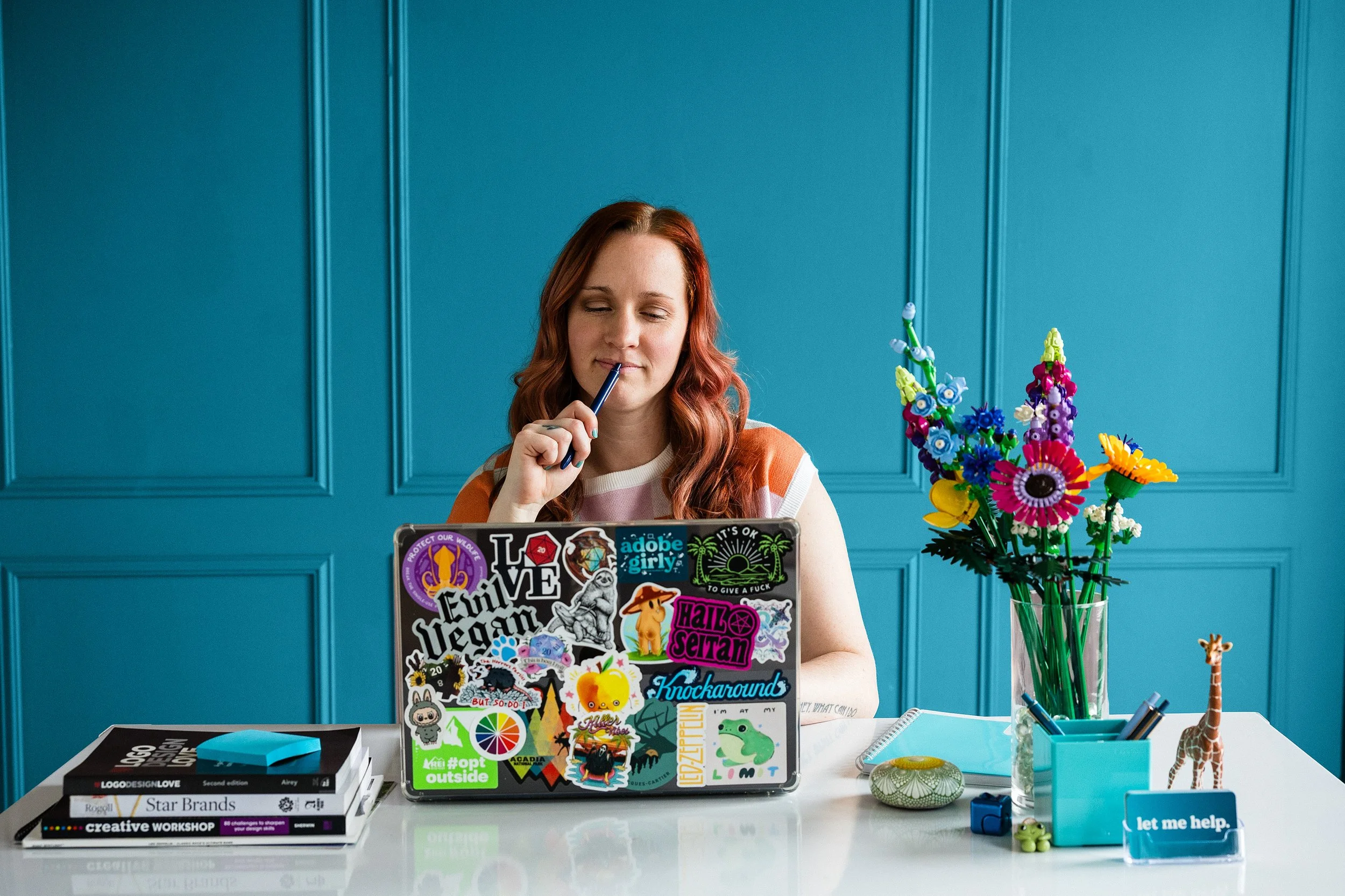 Kristen sitting at a white desk against a teal back drop while working on a laptop decorated with various stickers