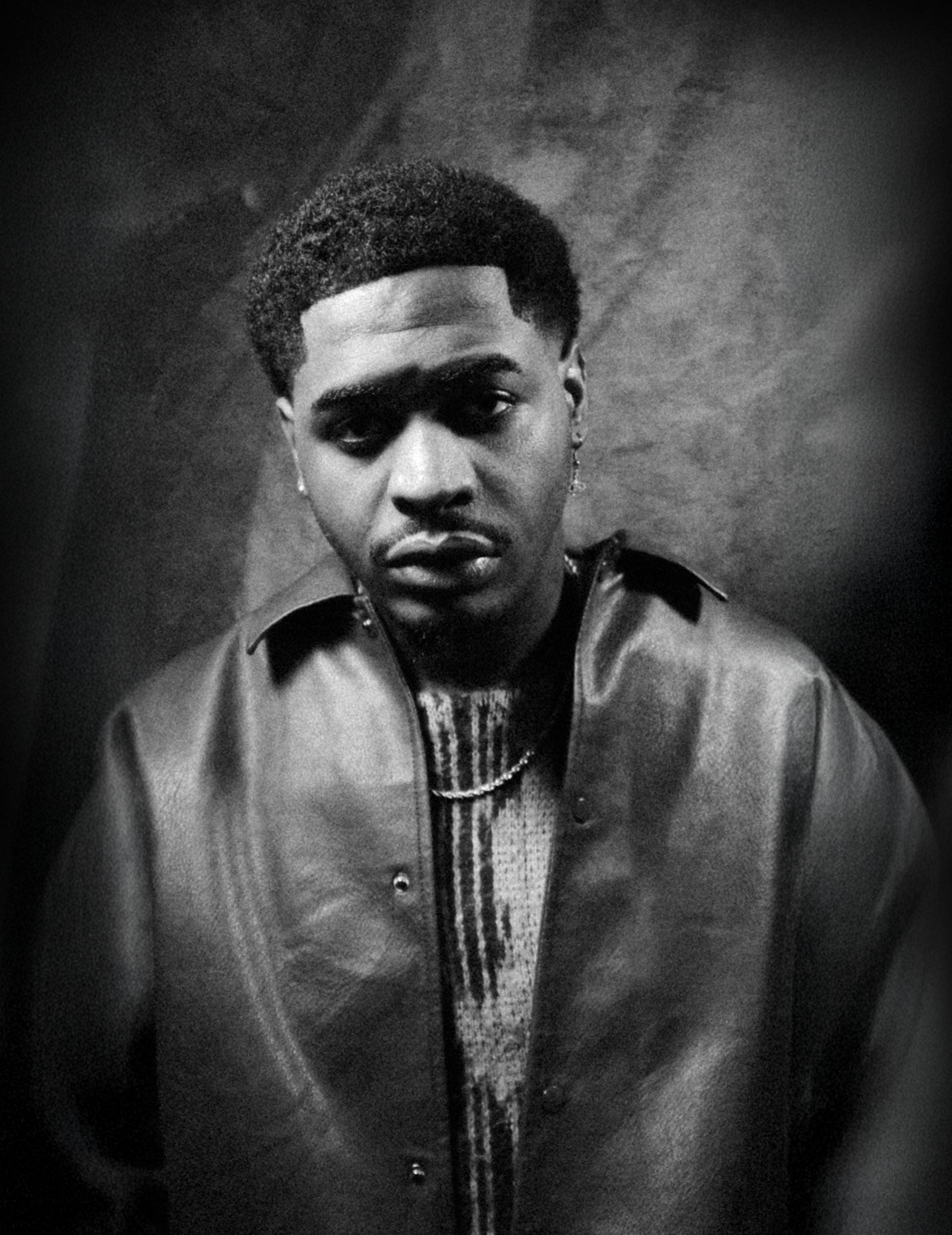 Black and white portrait of a Jah Croyant directly at the camera, wearing a leather jacket over a patterned sweater, with short hair and hoop earrings.