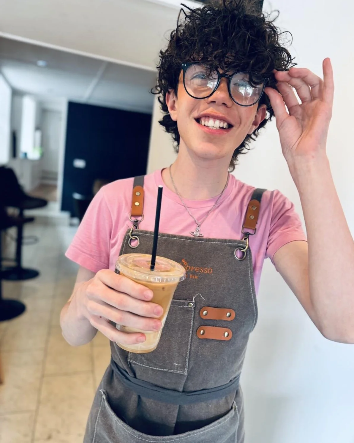 A cheerful person with curly hair and glasses holding an iced coffee and smiling in a coffee shop.