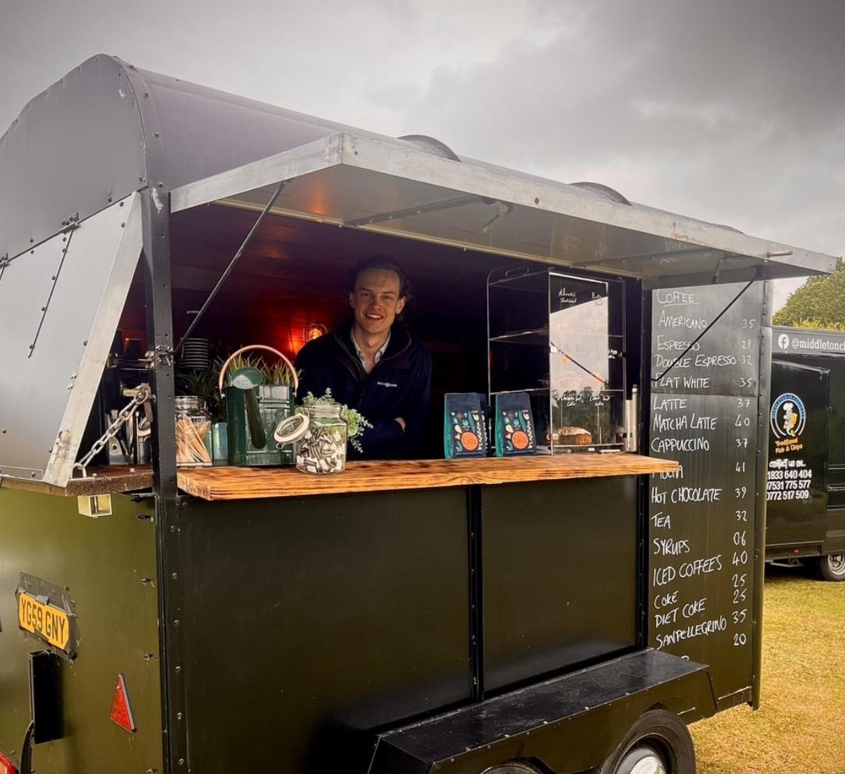 A man standing inside an outdoor coffee and snack trailer, smiling at the camera, with a menu board listing various drinks and prices.