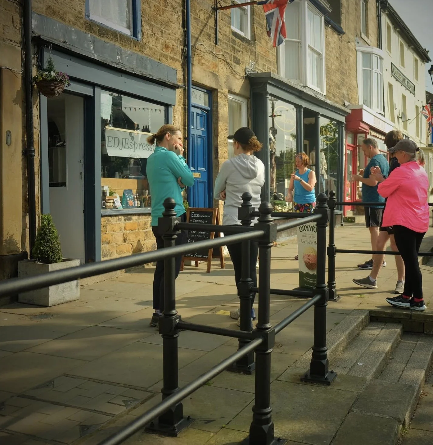 People standing in line outside a small coffee shop on a sidewalk with stone steps, black railings, and storefronts with colorful doors and windows, with flags hanging overhead.