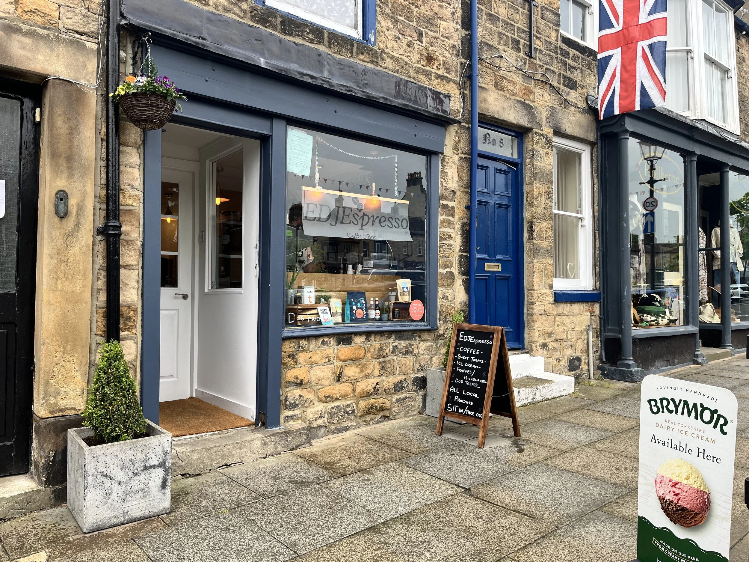 Exterior view of a small café named 'EDJ Espresso' with a large glass window and an entrance door, surrounded by brick walls. A chalkboard sign outside advertises coffee, sweet treats, ice cream, frappes, smoothies, and local produce. A decorative potted plant is next to the door. A signboard on the sidewalk promotes Brymor dairy ice cream, featuring an image of a scoop of ice cream in three flavors and a Union Jack flag is displayed on the building.