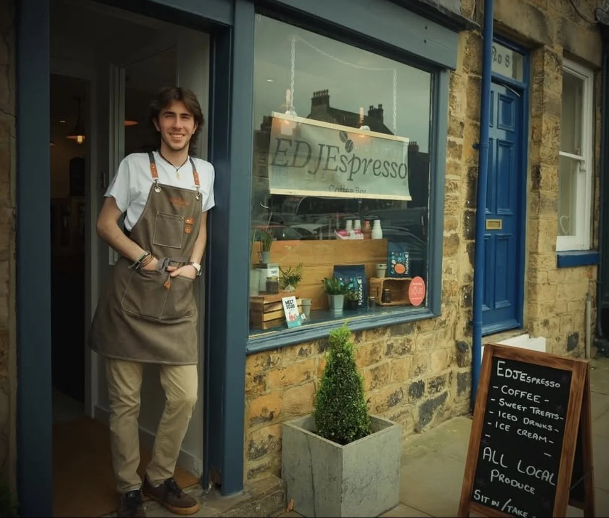 A young man with brown hair and a white shirt smiling, wearing a brown apron, standing at the entrance of a café called 'EDJ Espresso'. The café has a large window displaying plants and a sign, and a blackboard outside listing offerings like coffee, sweet treats, and ice cream.