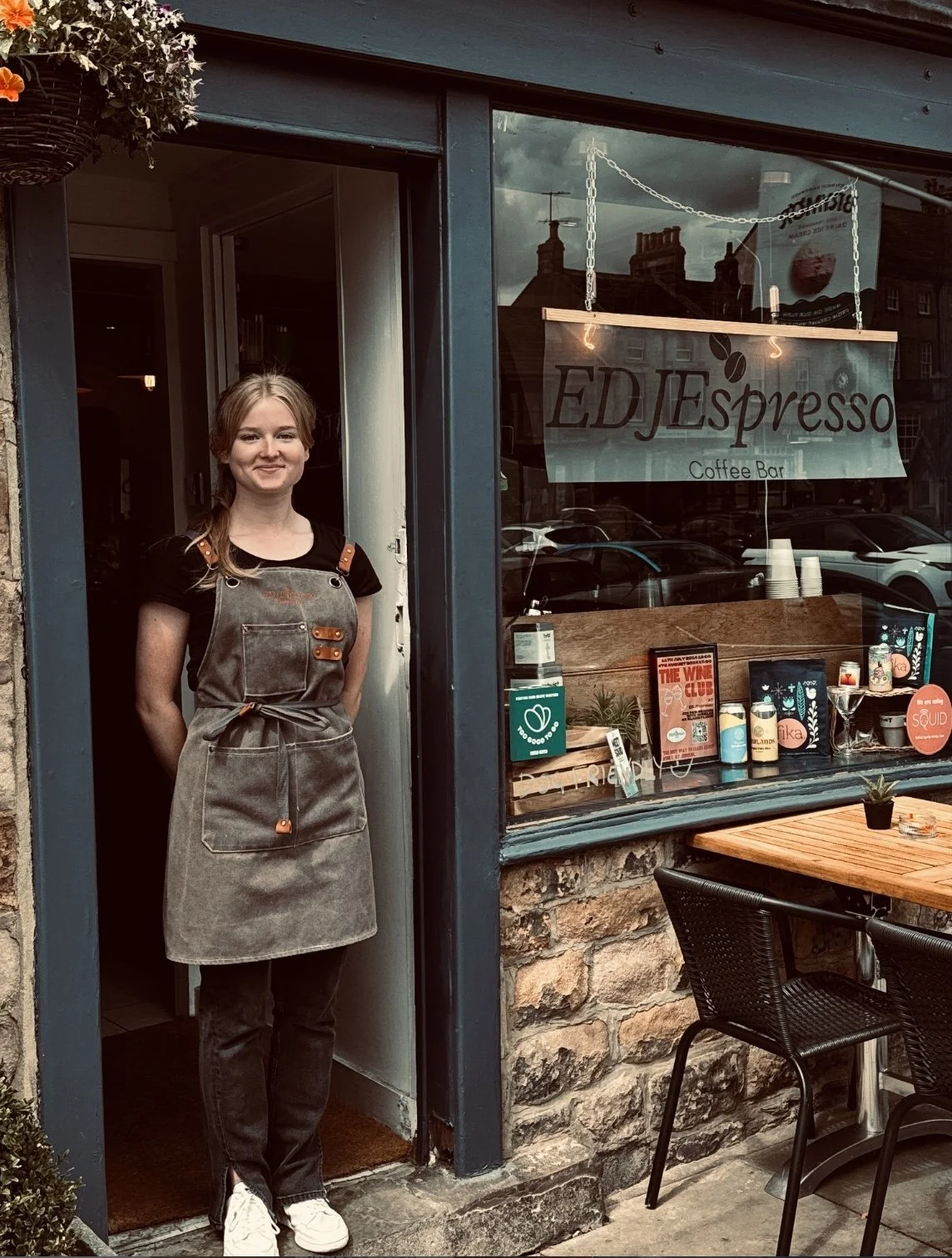 A young woman wearing a gray apron stands outside a café named 'EDJ Espresso' with a glass window displaying various items and promotional signs.