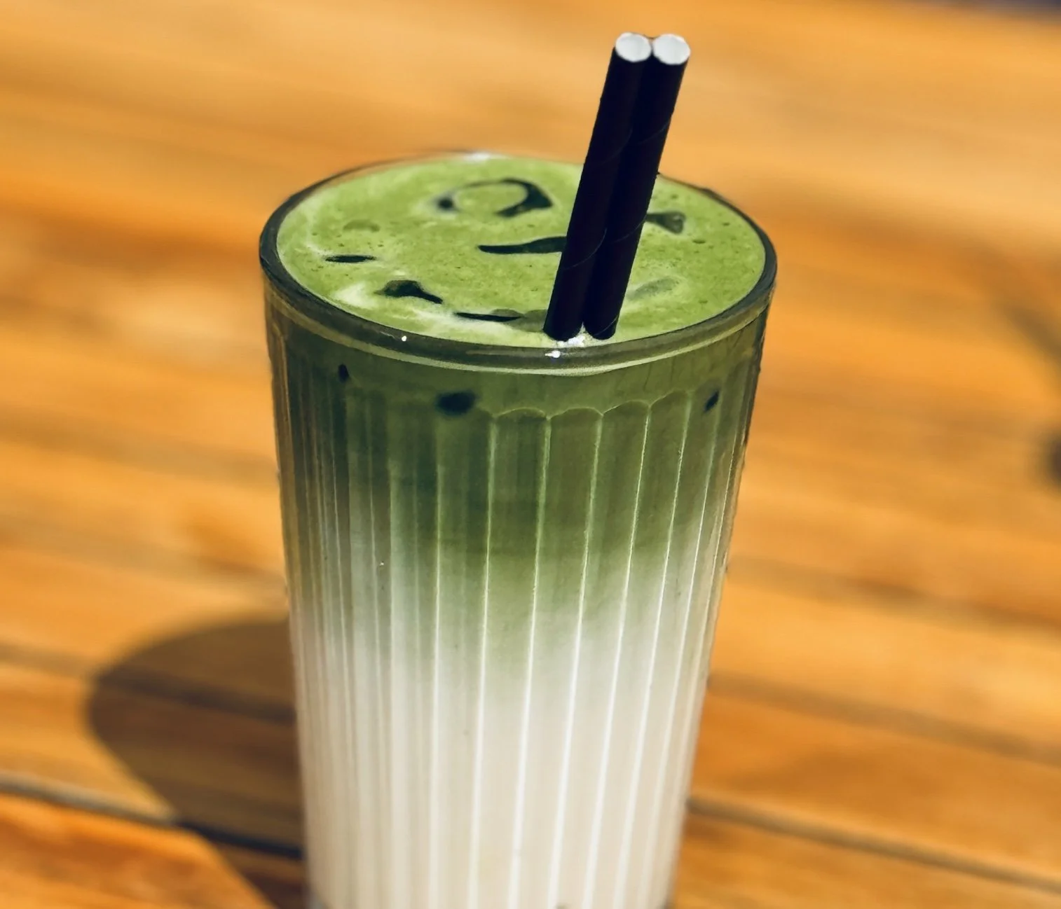 A tall glass of green smoothie with black straws on a wooden surface.