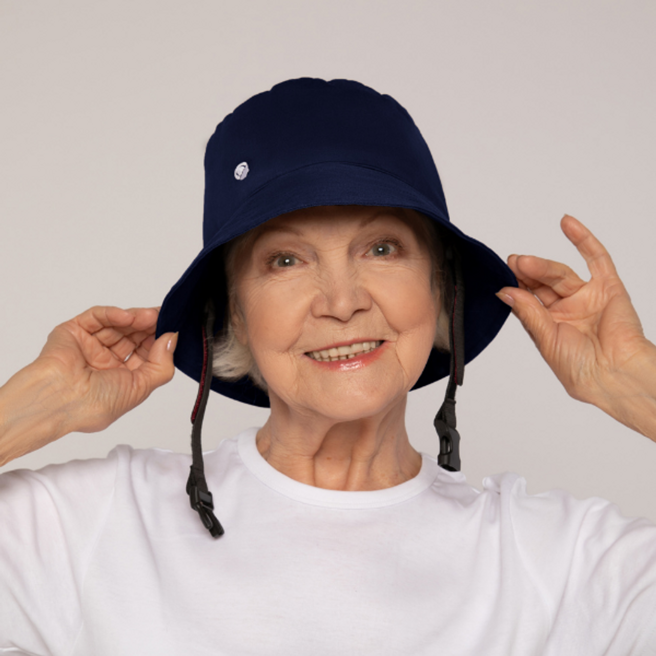 Elderly woman adjusting a navy blue bucket hat, wearing a white shirt, smiling against a plain background.