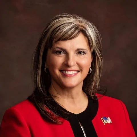 A woman with long, layered hair and highlights, wearing a red blazer with an American flag pin. She is smiling against a dark background.