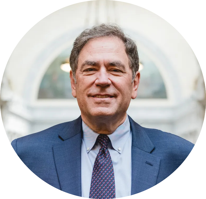A man in a blue suit and red patterned tie stands smiling in front of a blurred, arched background.