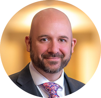 A bald man with a beard, wearing a suit and a colorful tie, smiles at the camera. The background is softly blurred, suggesting an indoor setting.