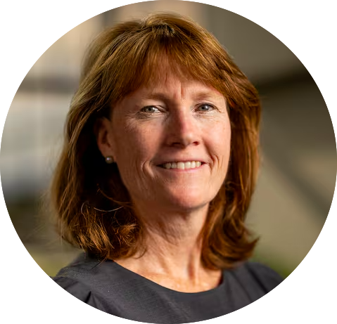 A woman with shoulder-length red hair, wearing a dark top, smiles at the camera. The background is softly blurred, emphasizing their face.