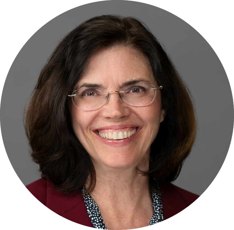 A woman with shoulder-length dark brown hair and glasses smiles warmly. They are wearing a dark blazer over a patterned blouse, set against a neutral gray background.