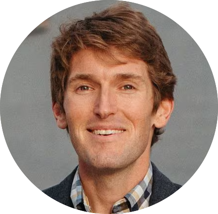 A man with short brown hair smiling, wearing a checkered shirt and a dark jacket. The background is neutral and blurred.