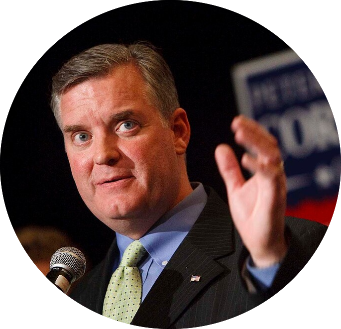 A man in a suit speaks into a microphone, his right hand raised. He wears a light green tie and has short hair. In the background, there are blurred elements with indistinct text and colors.