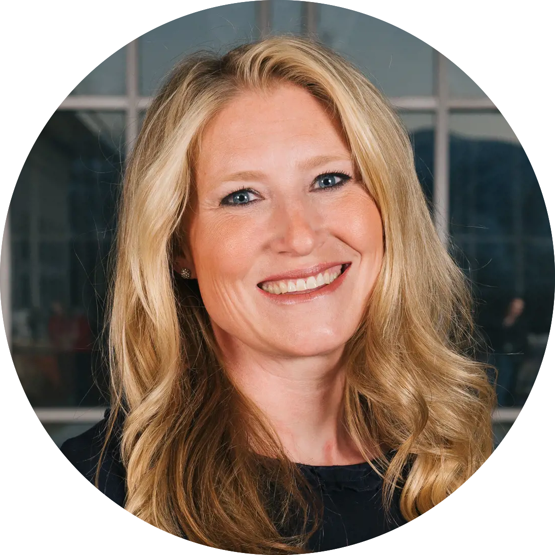 Smiling woman with long blonde hair and a dark top stands in front of a windowed building.