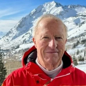 An man smiling while standing outdoors in a snowy mountain landscape. He is wearing a red jacket with a hood. The mountains and a clear blue sky are visible in the background.