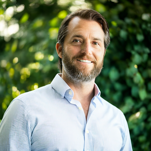 A man with a beard and light blue shirt smiles outdoors against a background of lush green foliage.
