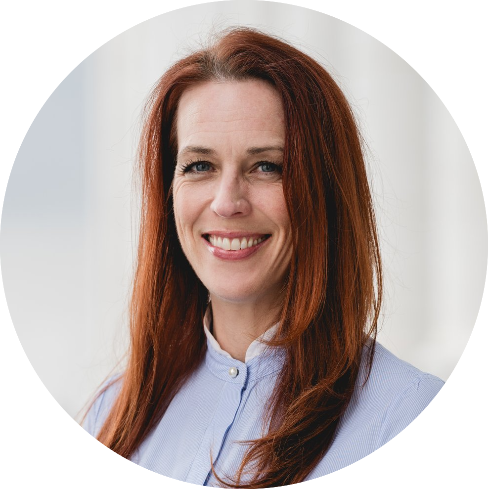 A woman with long red hair smiles at the camera. She is wearing a light blue collared shirt. The background is softly blurred, suggesting an indoor setting.