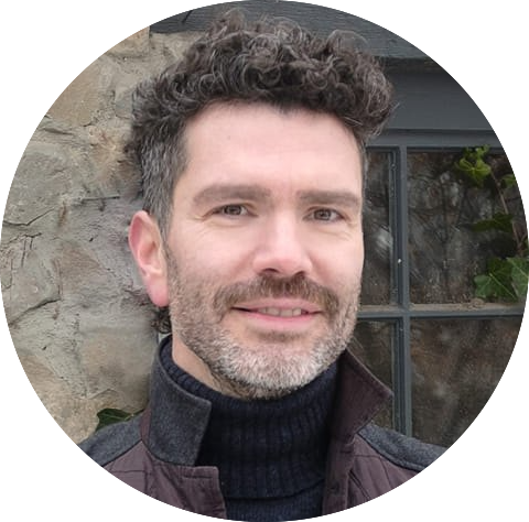 A man with curly hair and a beard is smiling. He is wearing a dark turtleneck sweater and a jacket, standing in front of a stone wall and window.