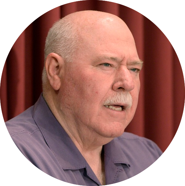 man with a white mustache and short hair speaks, wearing a purple collared shirt against a backdrop of deep red curtains.