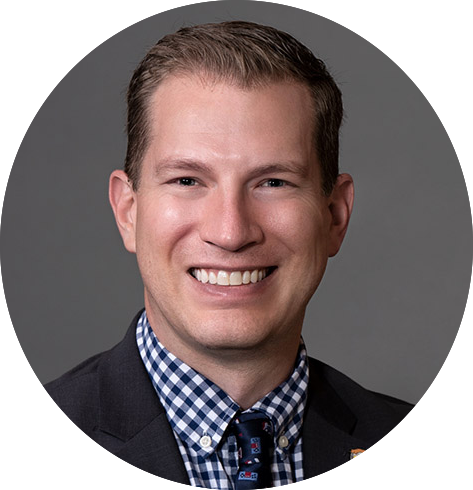 A man in a suit smiles at the camera. He is wearing a dark blazer, a checkered shirt, and a patterned tie. The background is a plain gray.