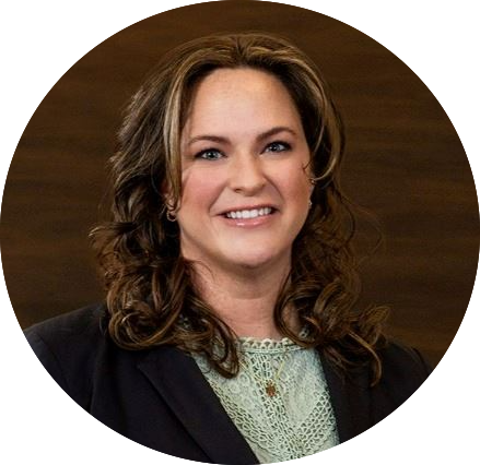 A woman with long, wavy brown hair smiles at the camera. She is wearing a dark blazer over a light green top, and the background is a plain brown wall.