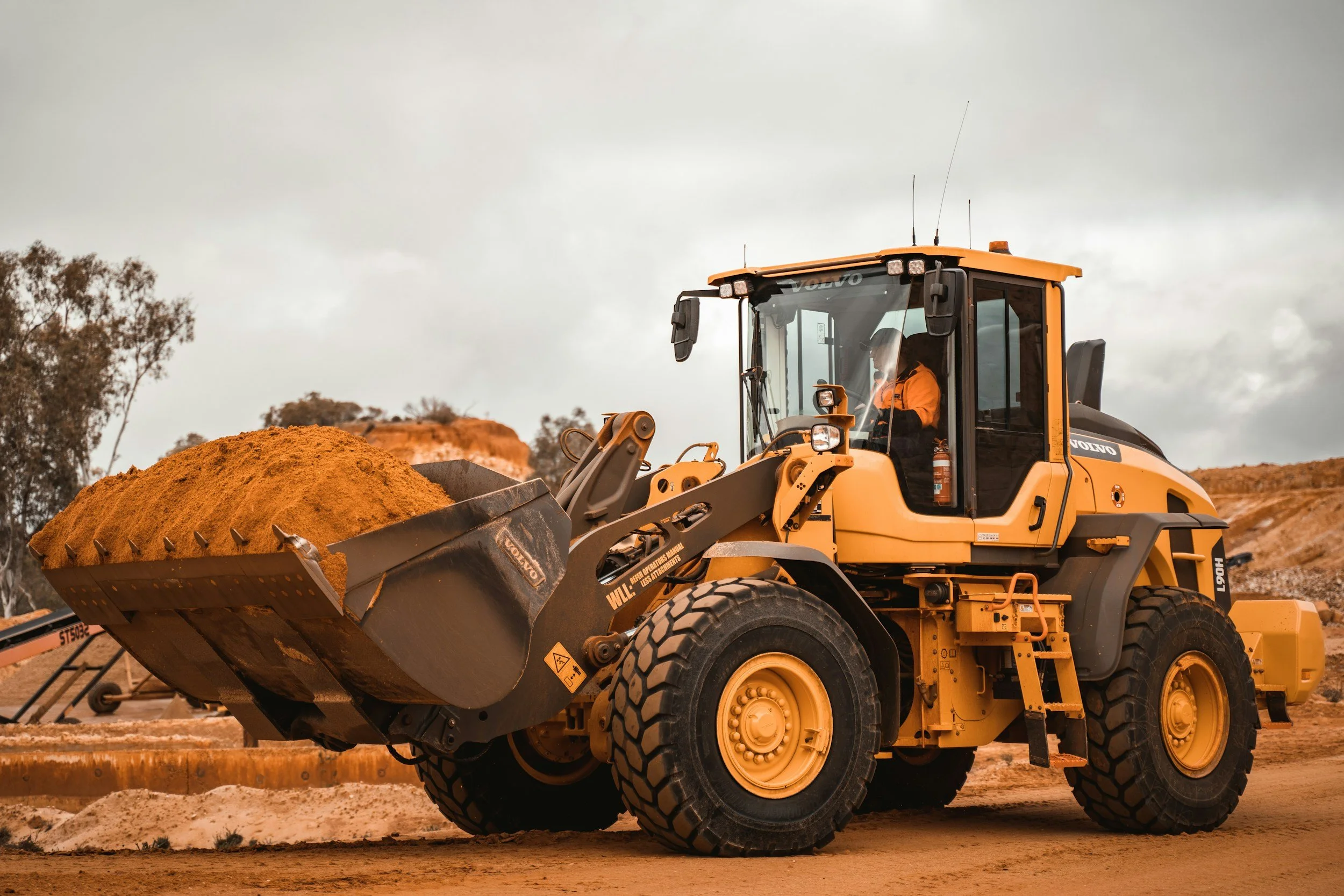 A yellow Volvo wheel loader carrying a load of dirt at a construction site on a cloudy day.