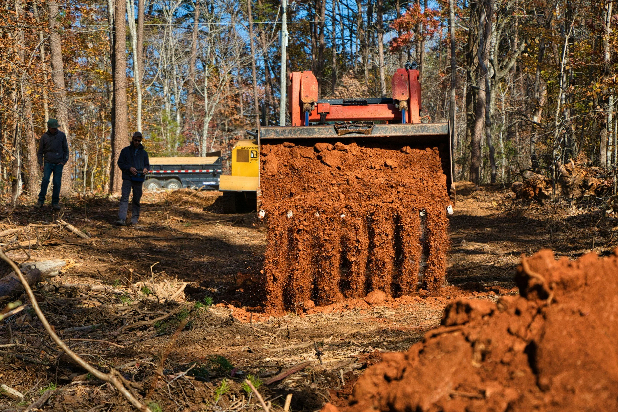 Tree removal equipment on a dirt path in a forest, with workers standing nearby and a trailer in the background.