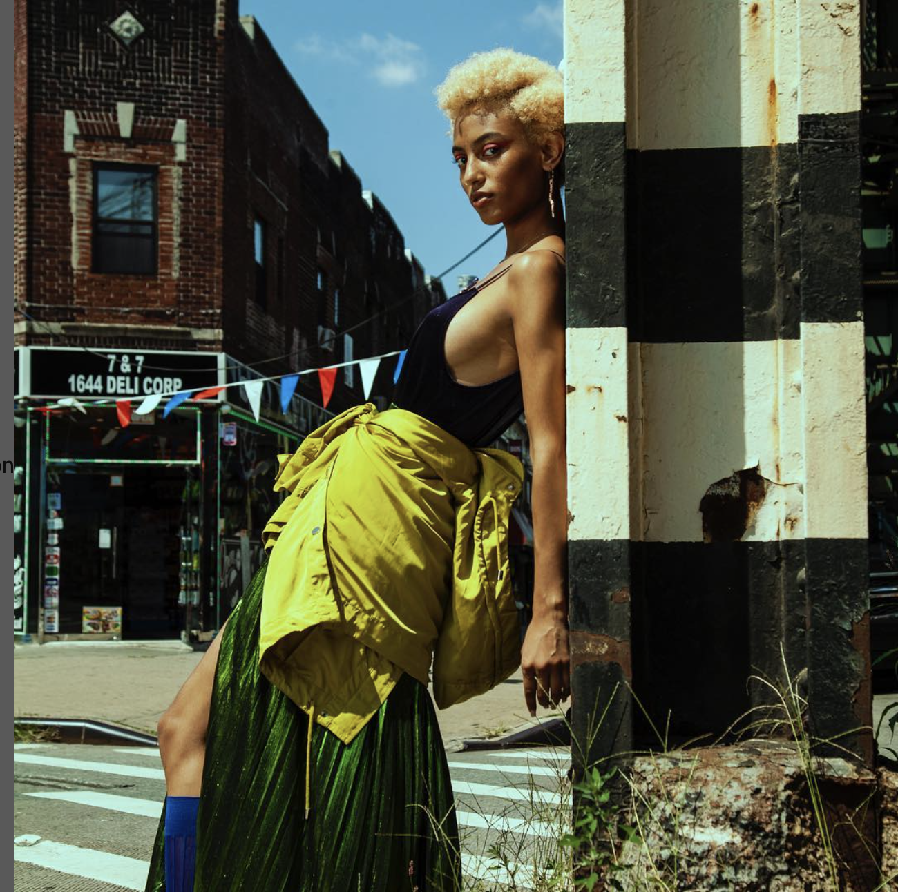 A woman with short, golden locks rests against a column adorned in black and cream stripes on an urban thoroughfare. She is dressed in a sleeveless black top paired with a flowing green pleated skirt, complemented by a yellow jacket casually knotted