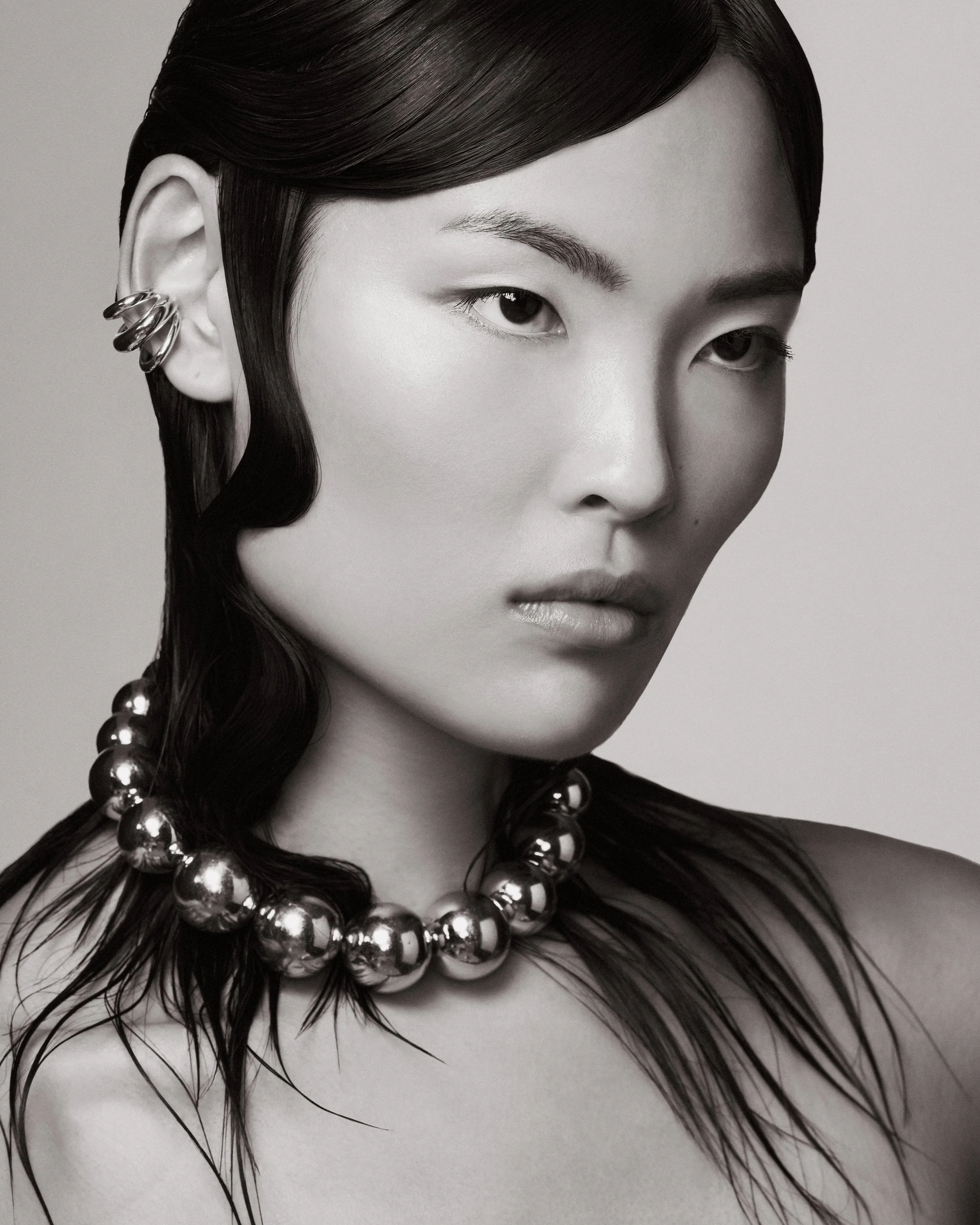 A black-and-white portrait of an East Asian woman with dark, wet hair styled in waves, wearing multiple earrings on her left ear, a chunky bead necklace, and minimal makeup, gazing slightly to the right.