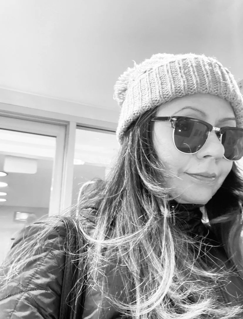 Woman wearing sunglasses and a knit beanie in a black-and-white photo, outdoors near a building with large windows.