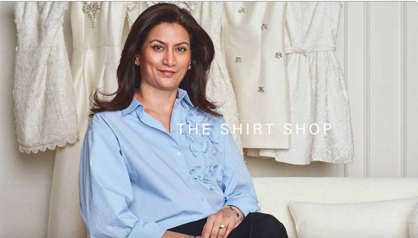 A woman is perched on a white couch, with white clothes hanging in the background. She's dressed in a light blue blouse adorned with floral embroidery, has dark brown hair, and is gazing at the camera. The text on the image says 'The Shirt Shop.'