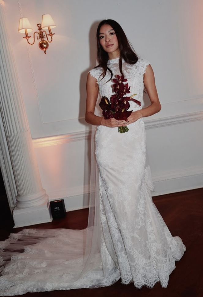A woman in a white lace wedding dress holding a bouquet of red flowers, standing indoors against a white wall with a brass wall sconce and a column, with a long veil trailing on the wooden floor.