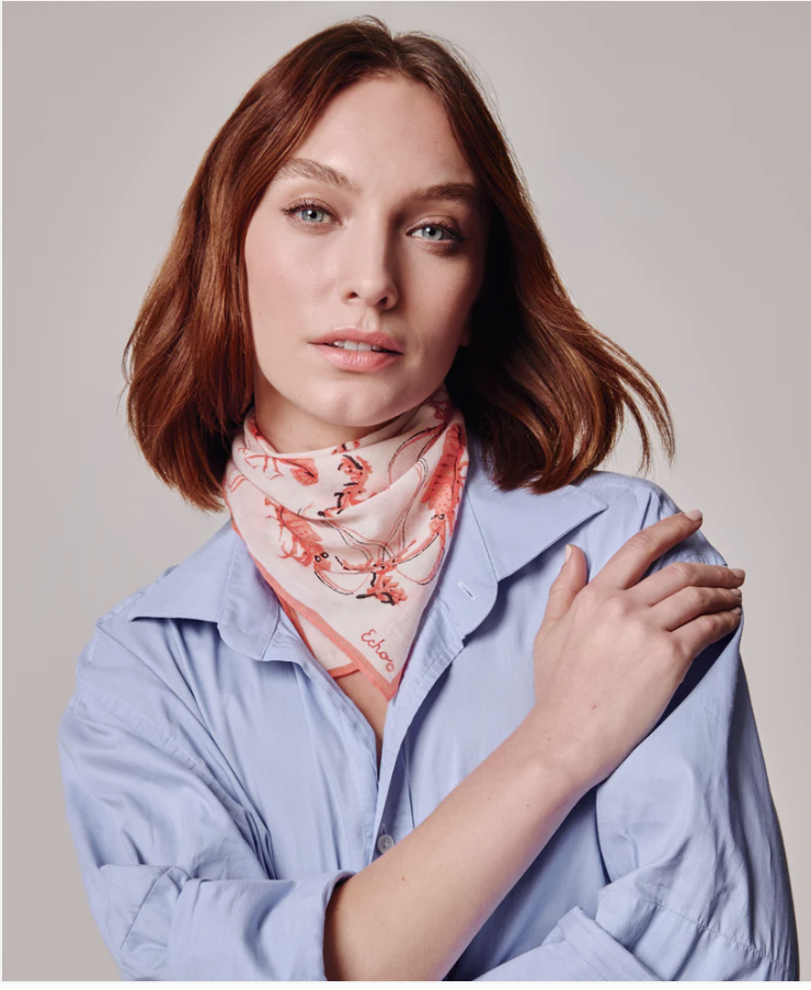 A woman with shoulder-length red hair, wearing a light blue shirt and a red and white scarf around her neck, looking at the camera with a relaxed expression.