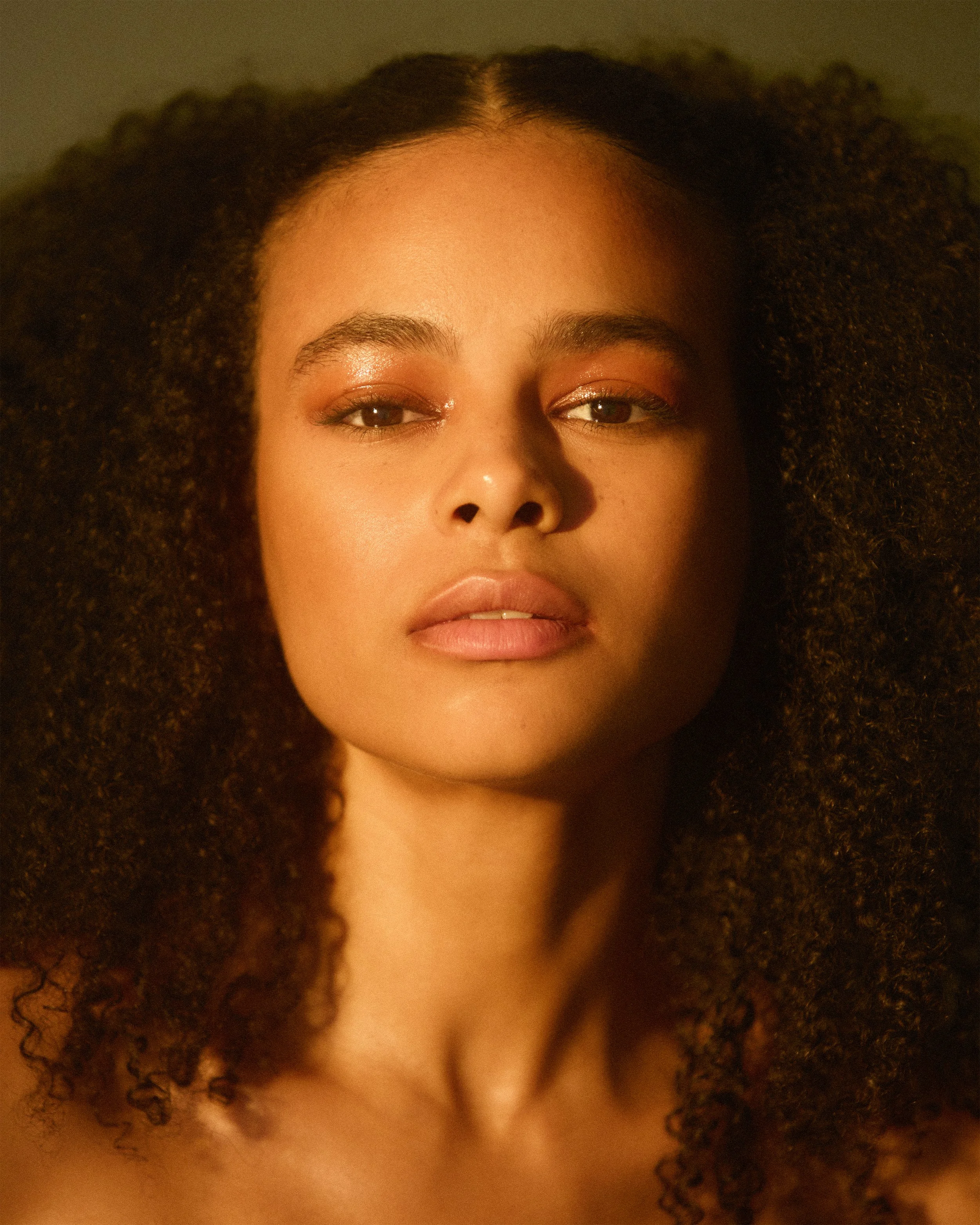 Close-up portrait of a woman with natural curly hair and light makeup, looking directly at the camera.