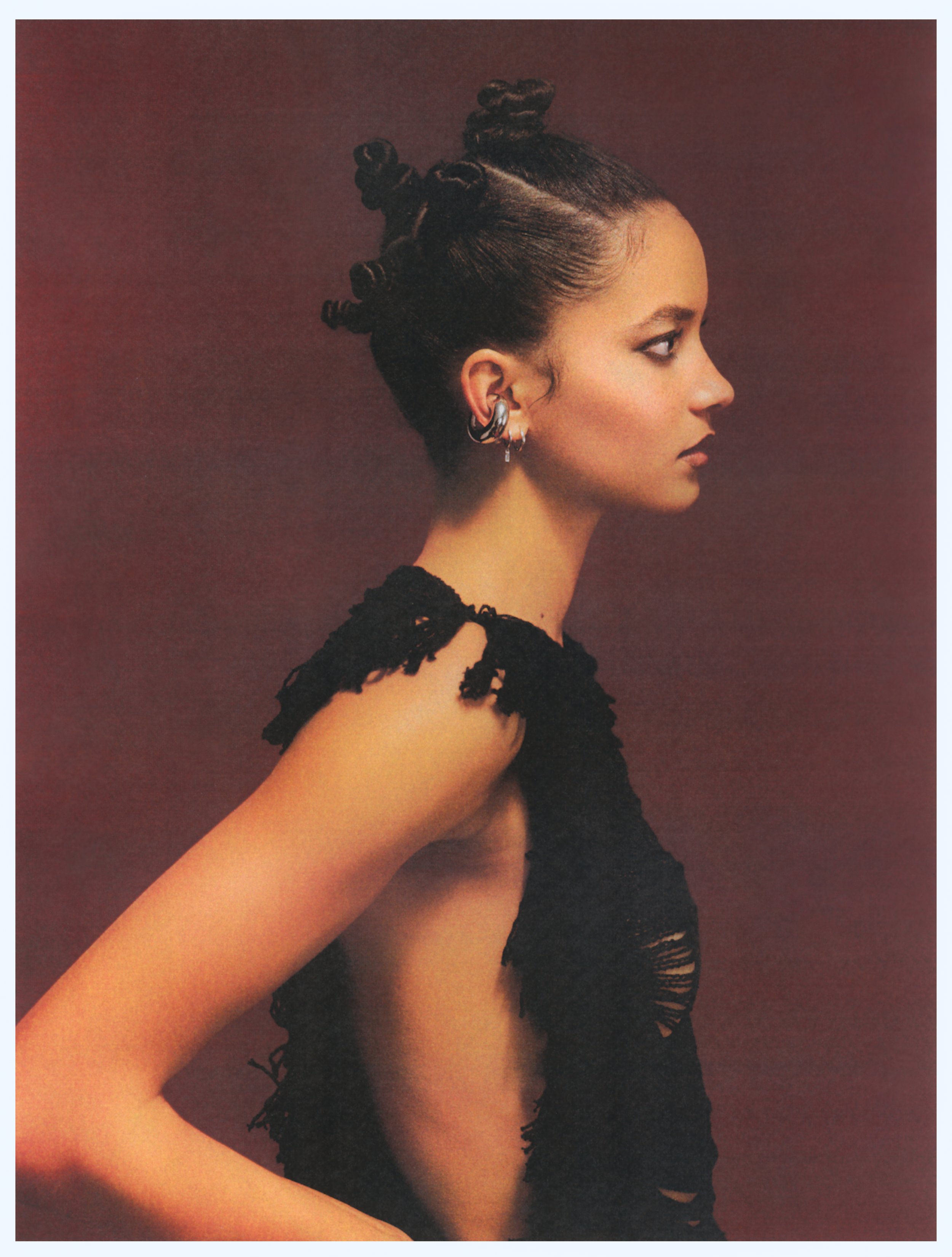 A side view of a woman showcasing her stylized hair and earrings, dressed in a black distressed top set against a dark red backdrop.