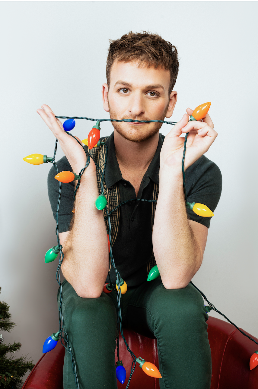 Young man sitting on a red chair, holding colorful Christmas lights with a playful expression.