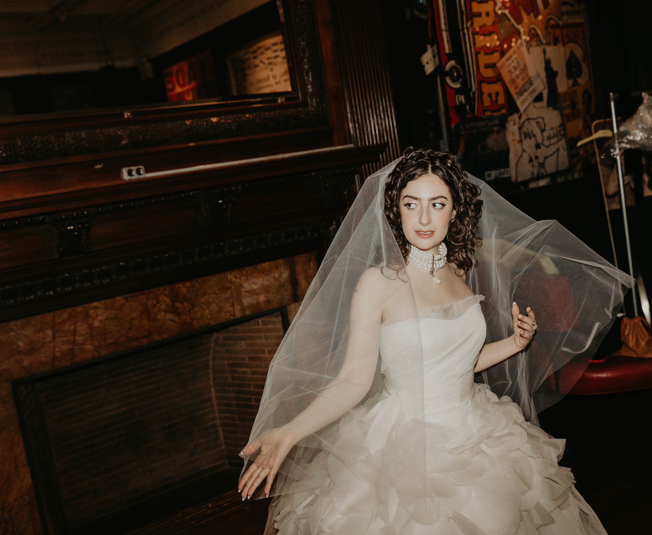A woman dressed in a wedding gown with a veil, standing indoors near a fireplace and a dark wooden wall with artistic decor. Famous comedian