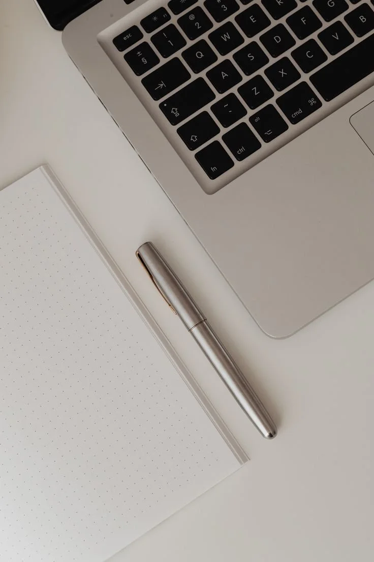 Close-up of a laptop keyboard, a silver pen, a dotted notebook, and a white surface.