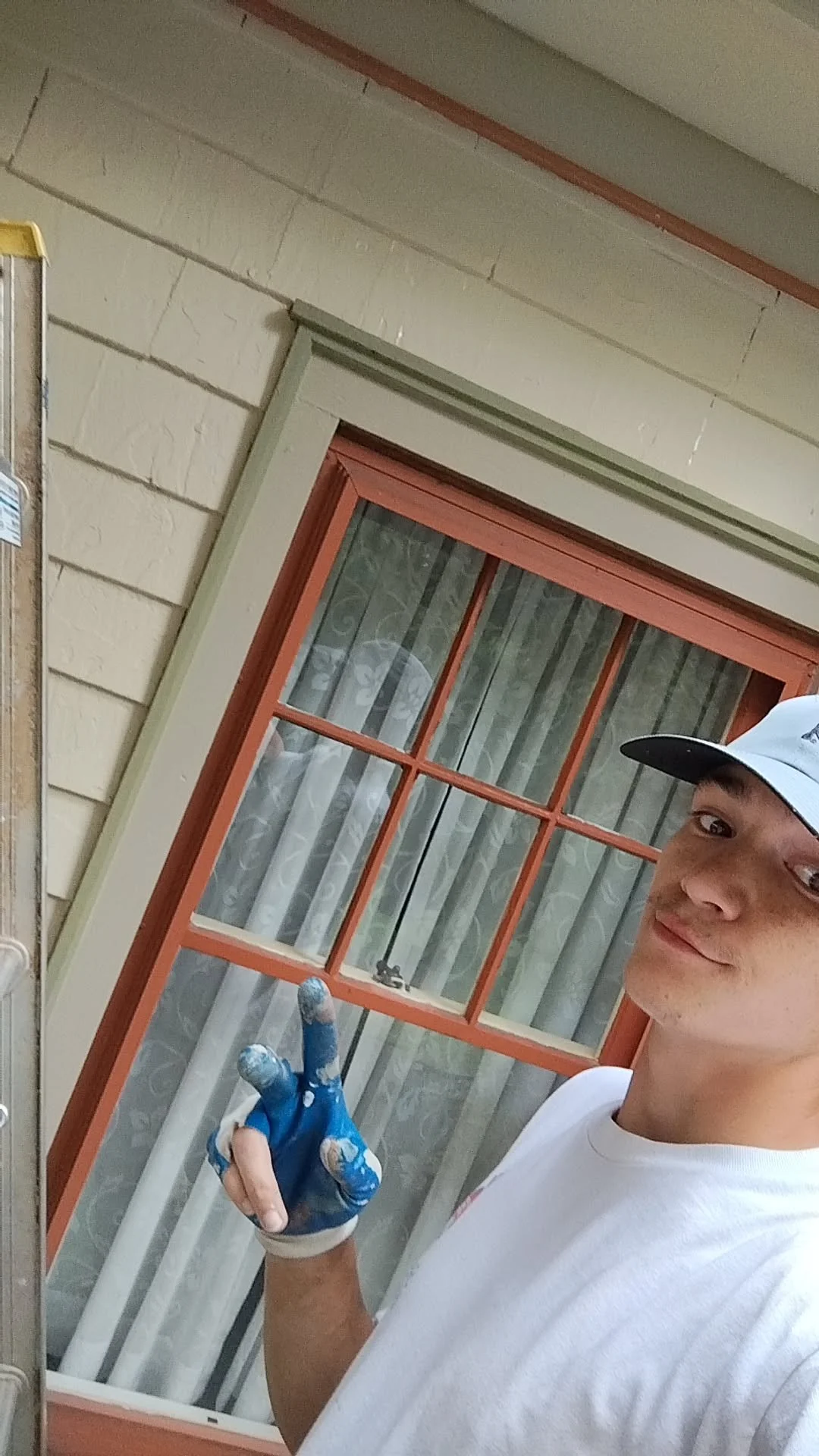 Young man wearing a white t-shirt and a white cap with a black brim, standing inside a store near a window with orange trim, giving a peace sign with his right hand covered in blue paint.