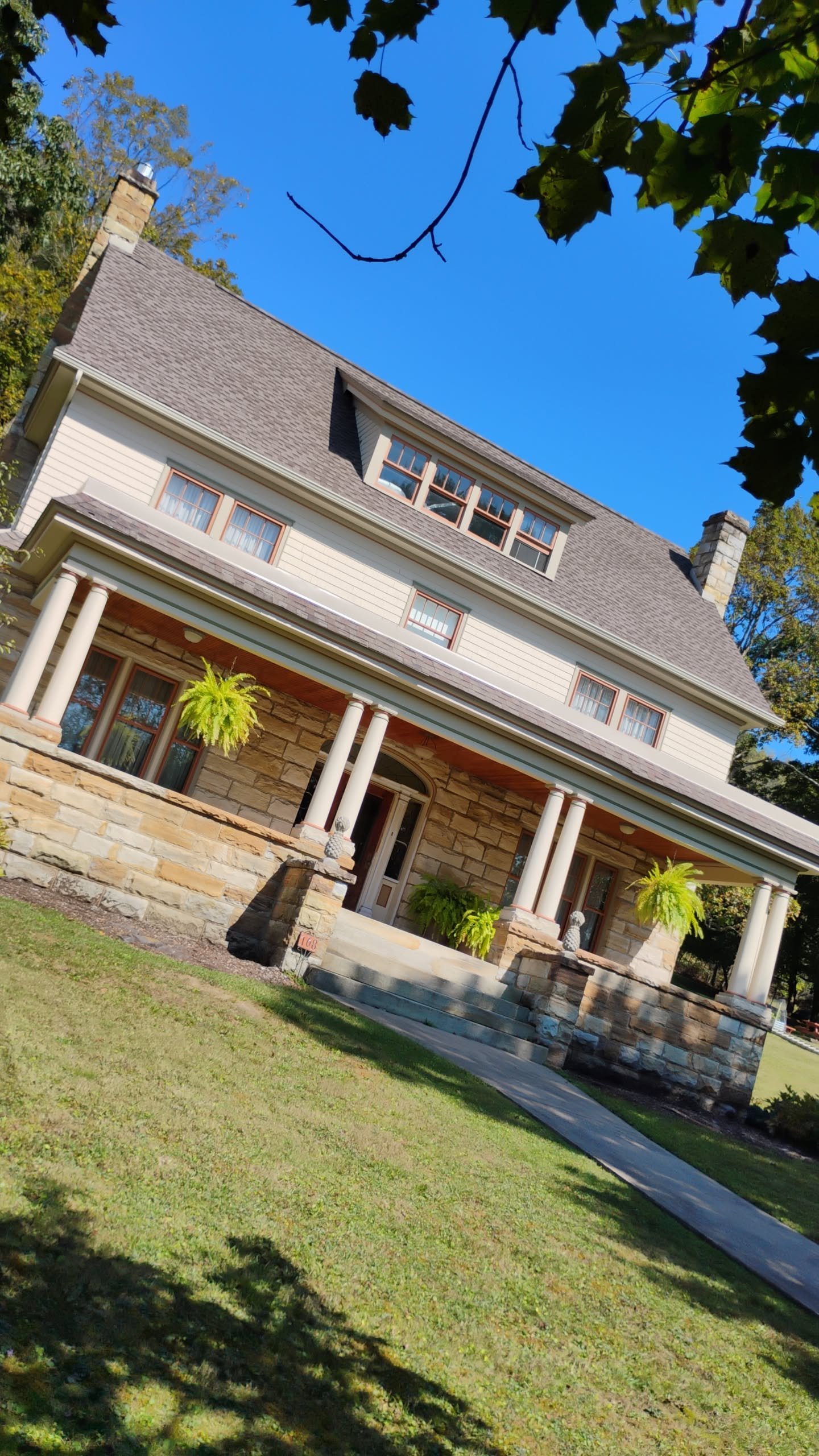 Front view of a large, multi-story house with a stone and siding exterior, a wide porch with columns, and a well-maintained lawn.