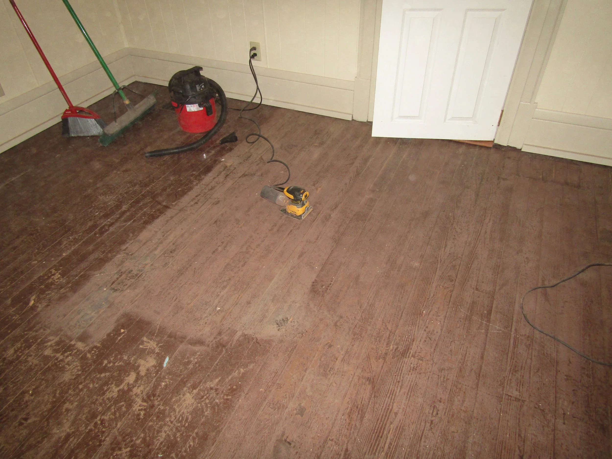 Room with hardwood floor being refinished, partial sanded with a sander on the floor, vacuum, broom, and dustpan visible in the corner.