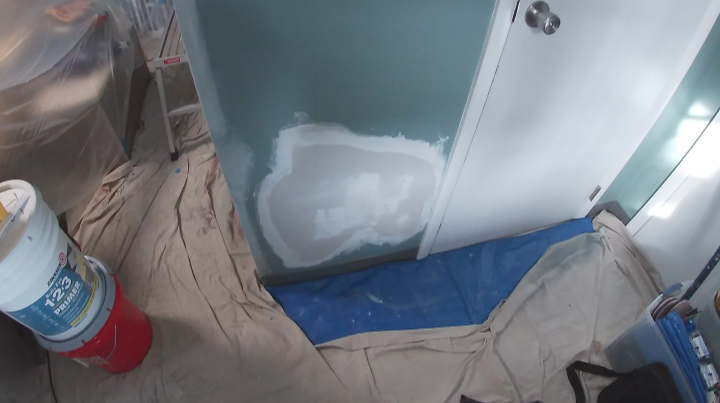 A partially painted wall with patch primer, drop cloths covering the floor, and painting supplies nearby.