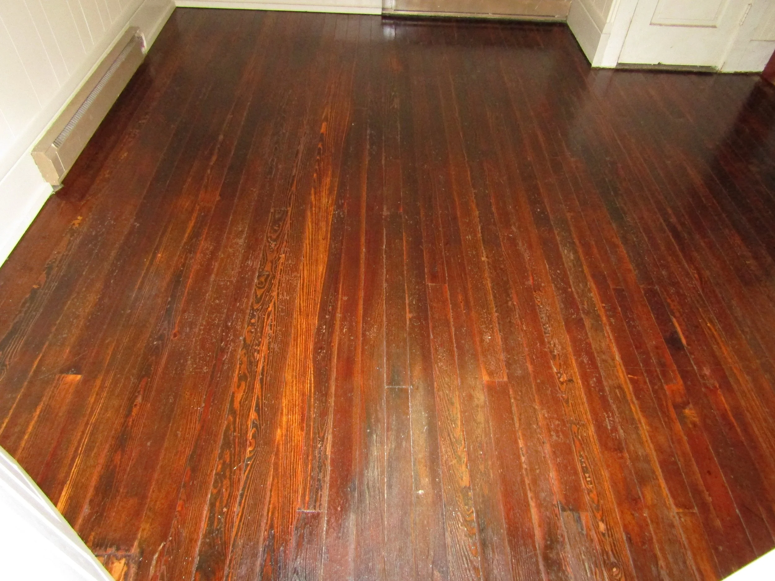 Finished hardwood floor with a dark reddish-brown stain and a polished finish in a room corner with white baseboards and a vent.